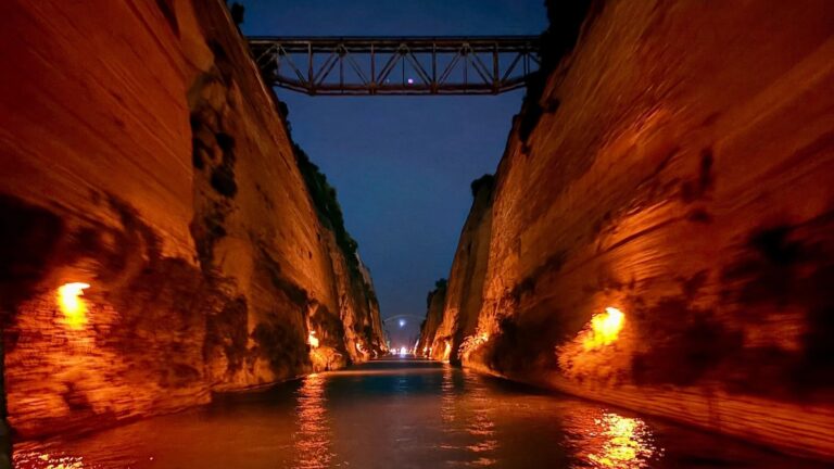 Foto notturna dello Stretto di Corinto vista dalla prua di una barca, con le alte pareti rocciose illuminate da luci calde, il ponte ferroviario in alto, la luna e i riflessi dorati sull'acqua scura del canale