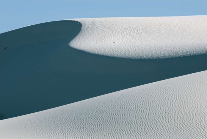 Primo piano astratto di dune di gesso candido a White Sands National Park, con increspature della sabbia e ombre profonde sotto un cielo azzurro chiaro.